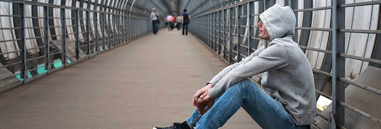 Jugendlicher in grauem Hoodie hockt an einem Brückengelände