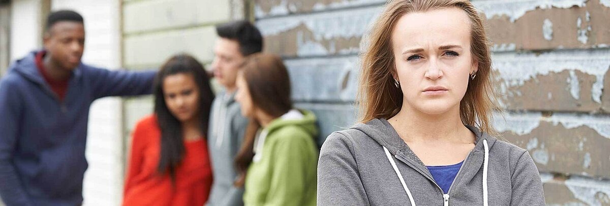 Jugendliche in grauem Hoodie blickt ernst in die Kamera, im Hintergrund eine diverse Gruppe weiterer Jugendlicher