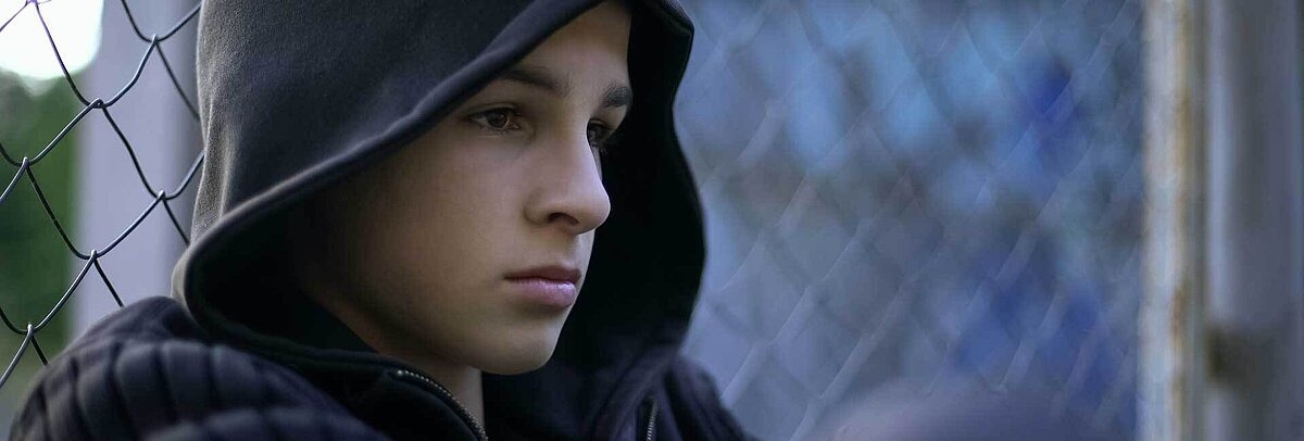 Nahaufnahme eines Jungen in blauem Hoodie, der vor einem Maschendrahtzaun hockt