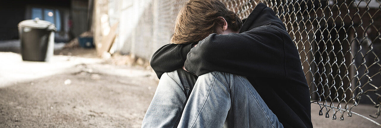 Teenager in schwarzem Hoodie hockt vor Maschendrahtzaun