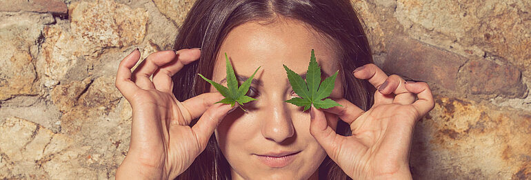 Nahaufnahme vn Mädchen, das sich zwei Cannabisblätter vor die Augen hält