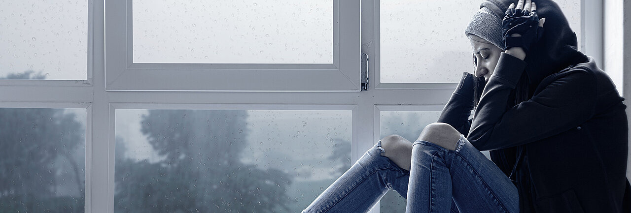 Teenagerin sitzt auf Fensterbank und hält sich die Ohren zu