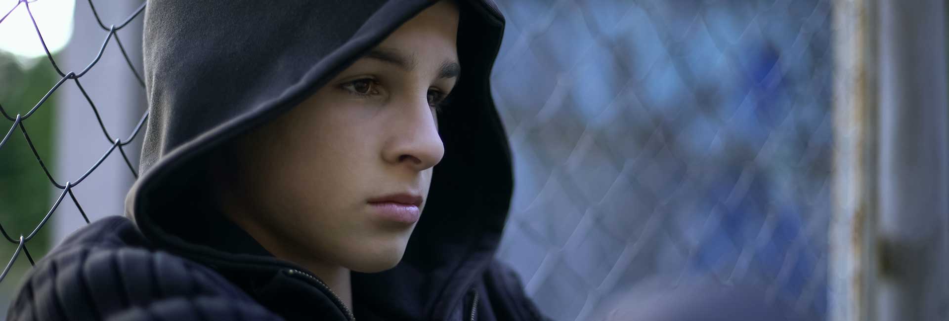 Nahaufnahme eines Jungen in blauem Hoodie, der vor einem Maschendrahtzaun hockt
